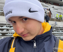 Close-up portrait of a person in a white beanie and winter jacket, standing in snowy stadium bleachers.